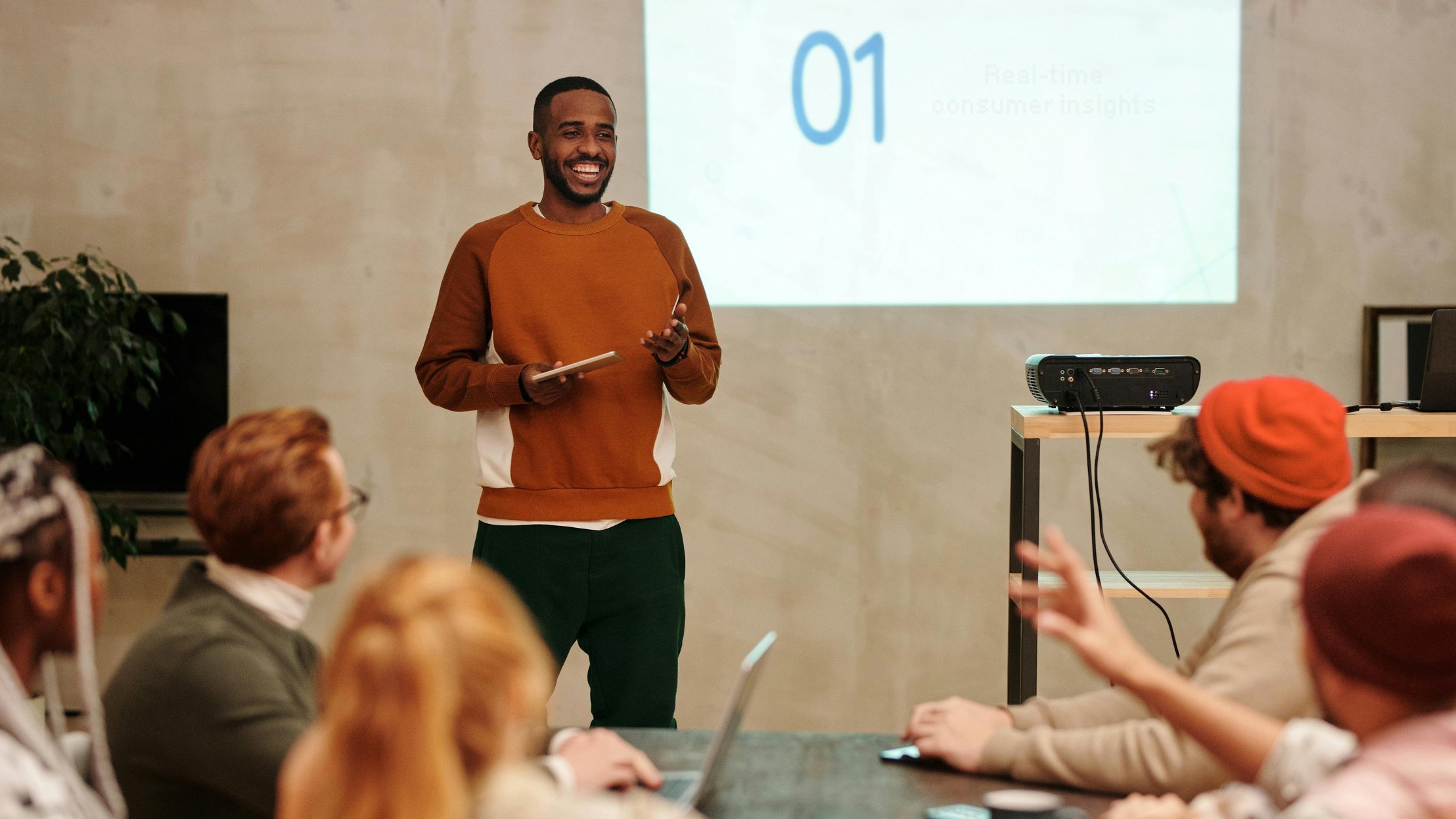 A presenter wearing a casual sweater holds a tablet and smiles while speaking to a group of diverse individuals seated at a table, with a projected slide titled "01 Real-time consumer insights" in the background.