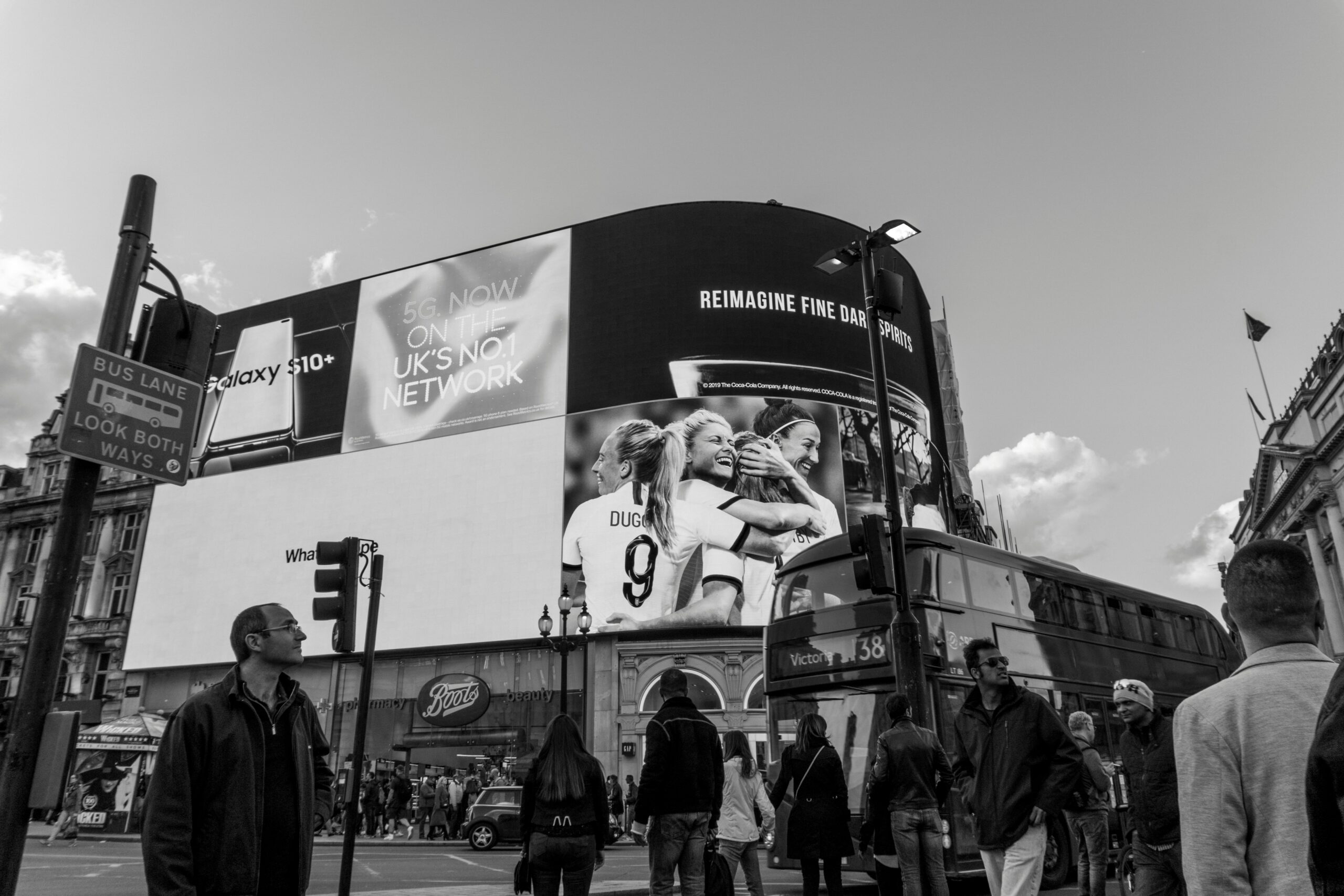 Piccadilly Circus London with digital billboards, Samsung Galaxy S10+ ad, 5G network sign, female footballers celebrating, double-decker bus, and pedestrians crossing.